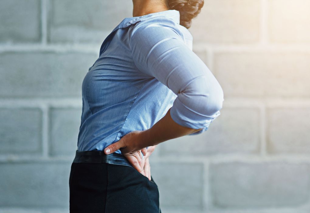 Frau greift sich im Stehen an den unteren Rücken vor neutralem Hintergrund.